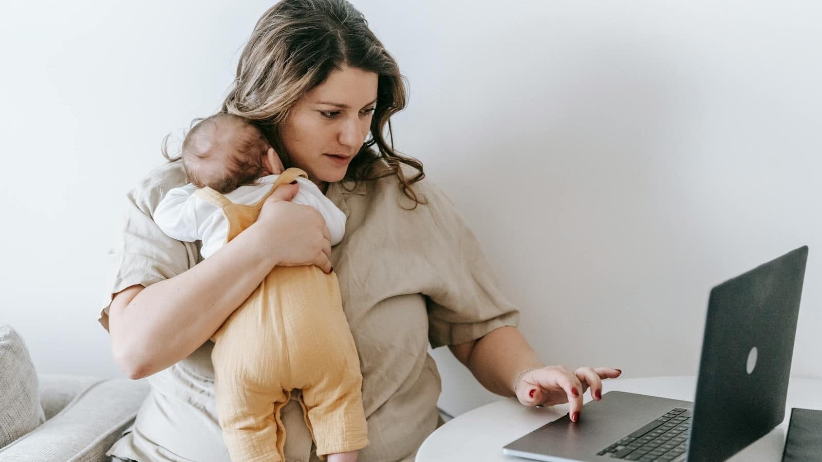 Concentrated young female freelancer embracing newborn while sitting at table and working remotely on laptop at home