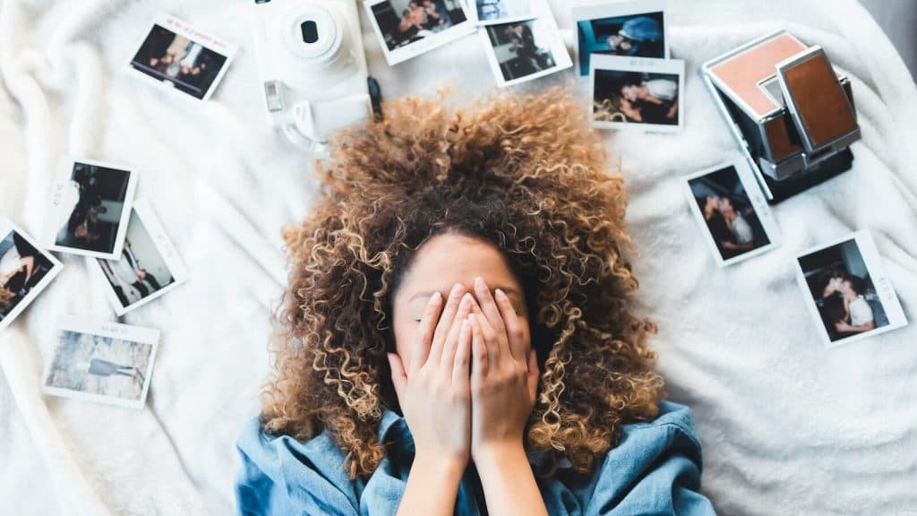woman lying on bed covering her face surrounded by photos and white camera